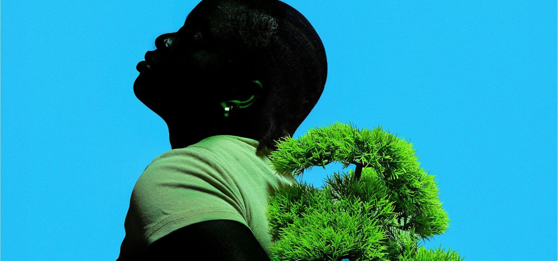 A black figure against a blue background. Wearing green and posing with natural foliage. 