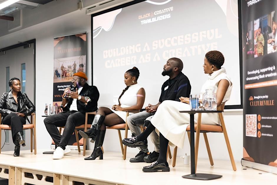 A panel of black creatives sit on a stage in discussion. The branding for Black Creative Trailblazers is displayed behind them. 
