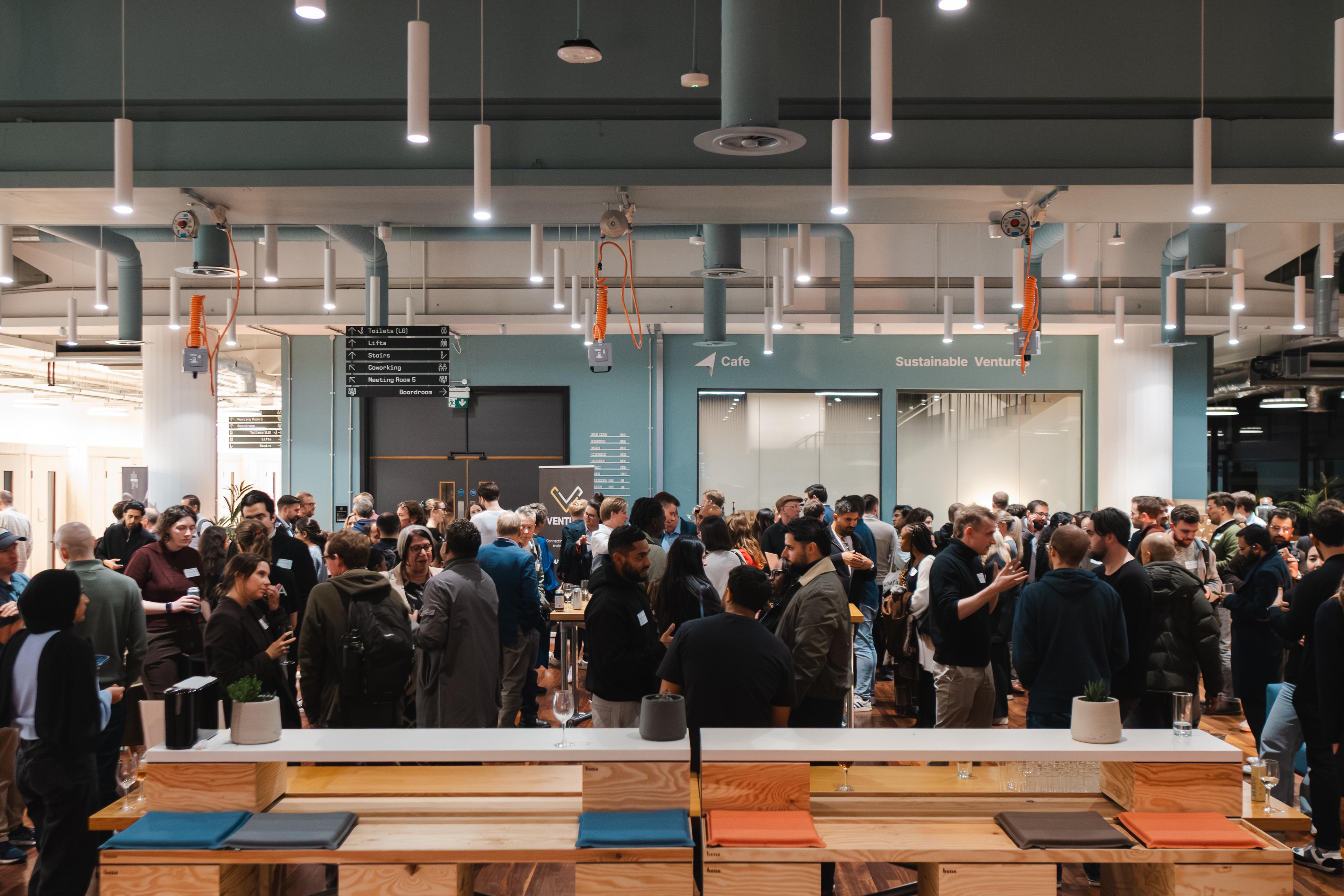 A diverse crowd of business professionals gather in a large café space chatting and socialising with one another. 